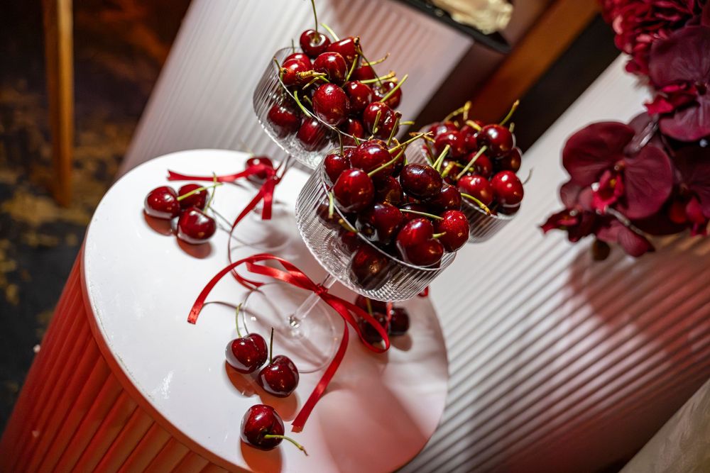 Fresh red cherries displayed in a glass bowl on a white tiered stand, decorated with red ribbon accents.