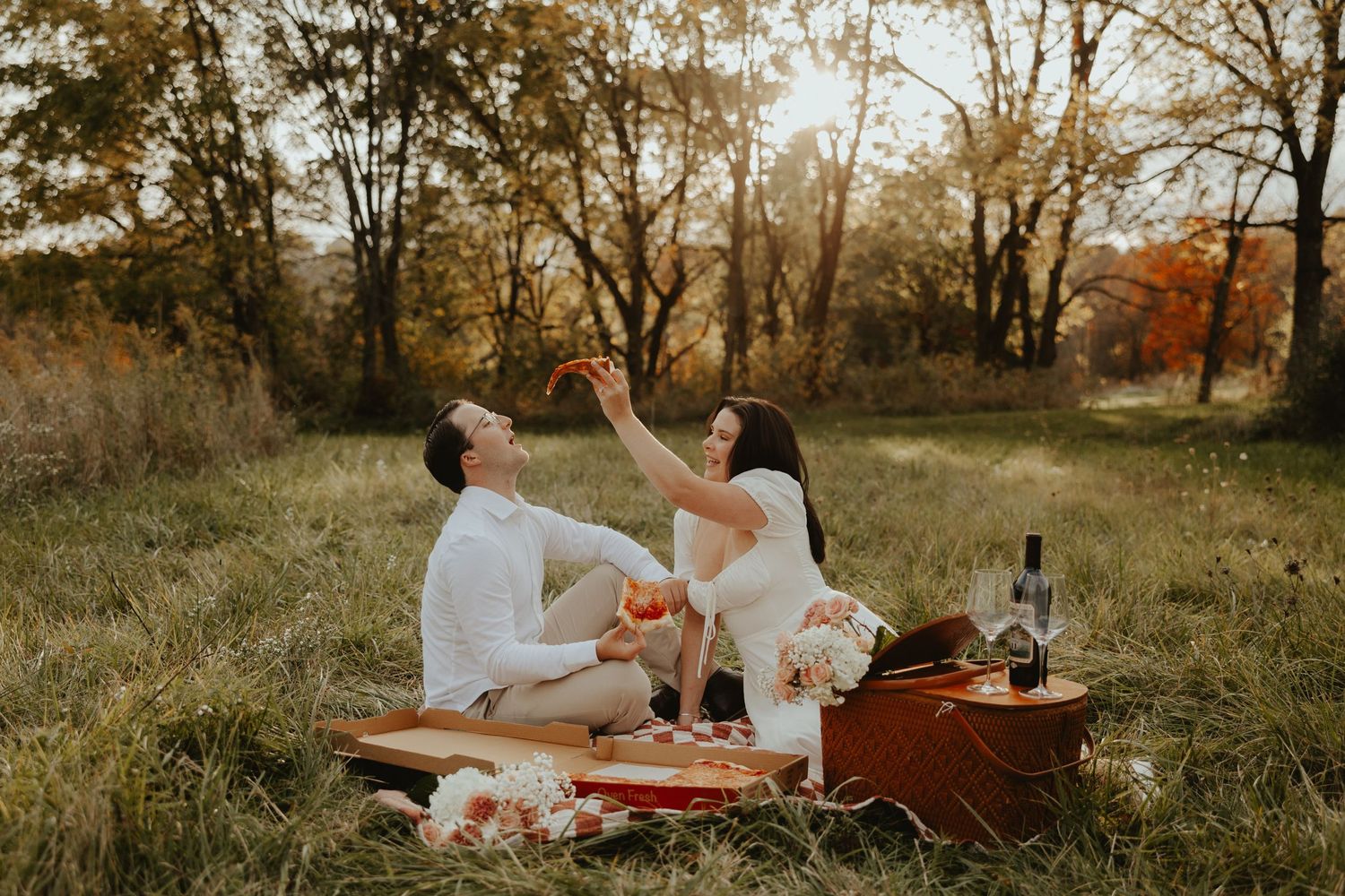Couple sharing pizza and wine at golden hour picnic on checkered blanket in autumn field.