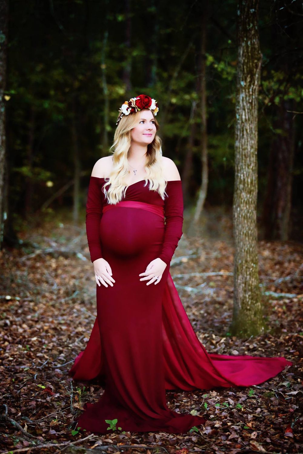 A maternity photo featuring a burgundy off-shoulder gown and floral crown in a forest setting.