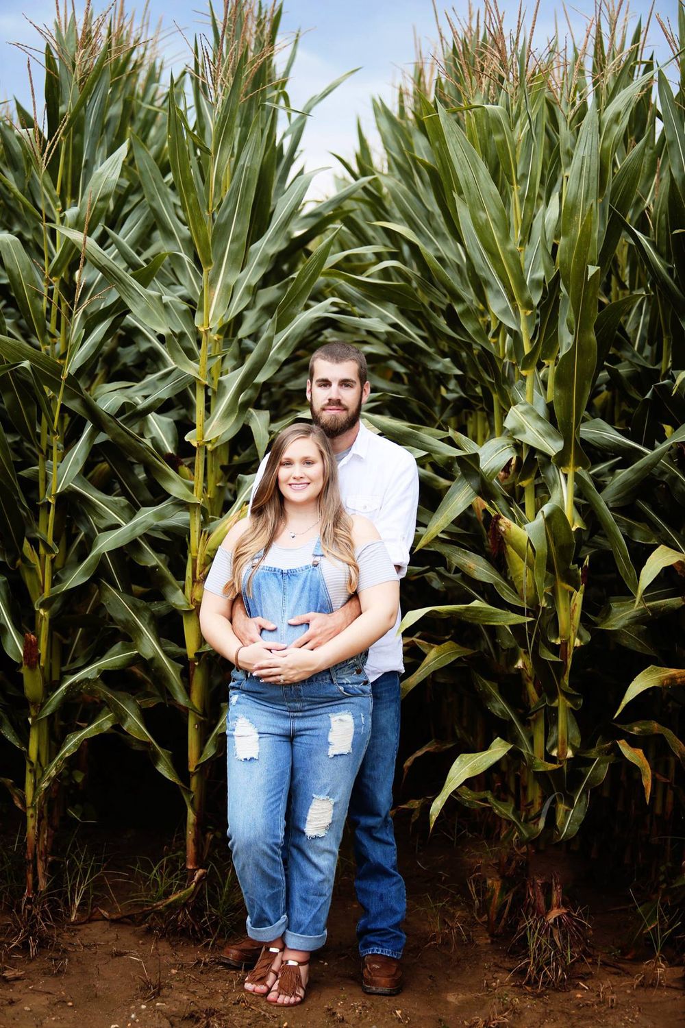 Couple poses in front of tall cornfield stalks on a bright summer day.