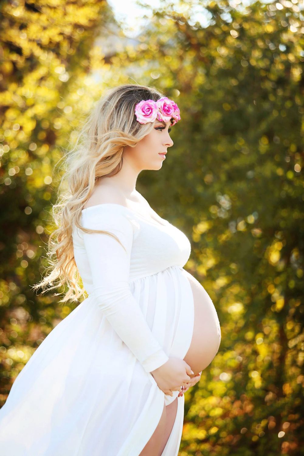 Ethereal maternity photo of woman in white dress with pink flower crown against golden sunlit background.