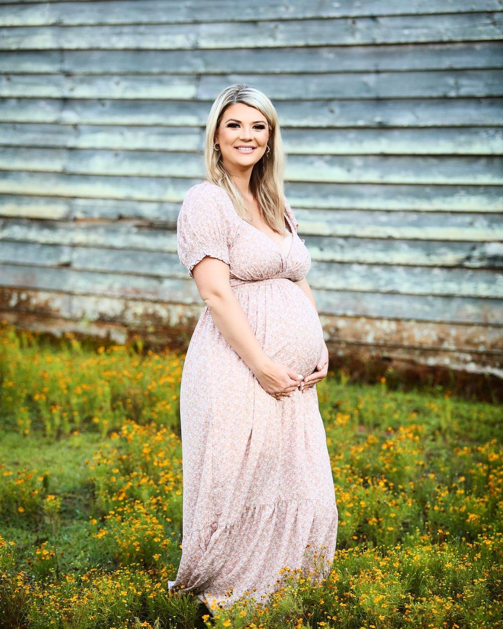 Maternity photo of woman in pink lace dress standing in field of wildflowers.