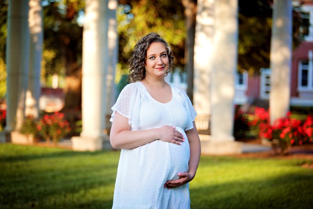A maternity portrait featuring a woman in white dress standing in a garden with pillars and red flowers.