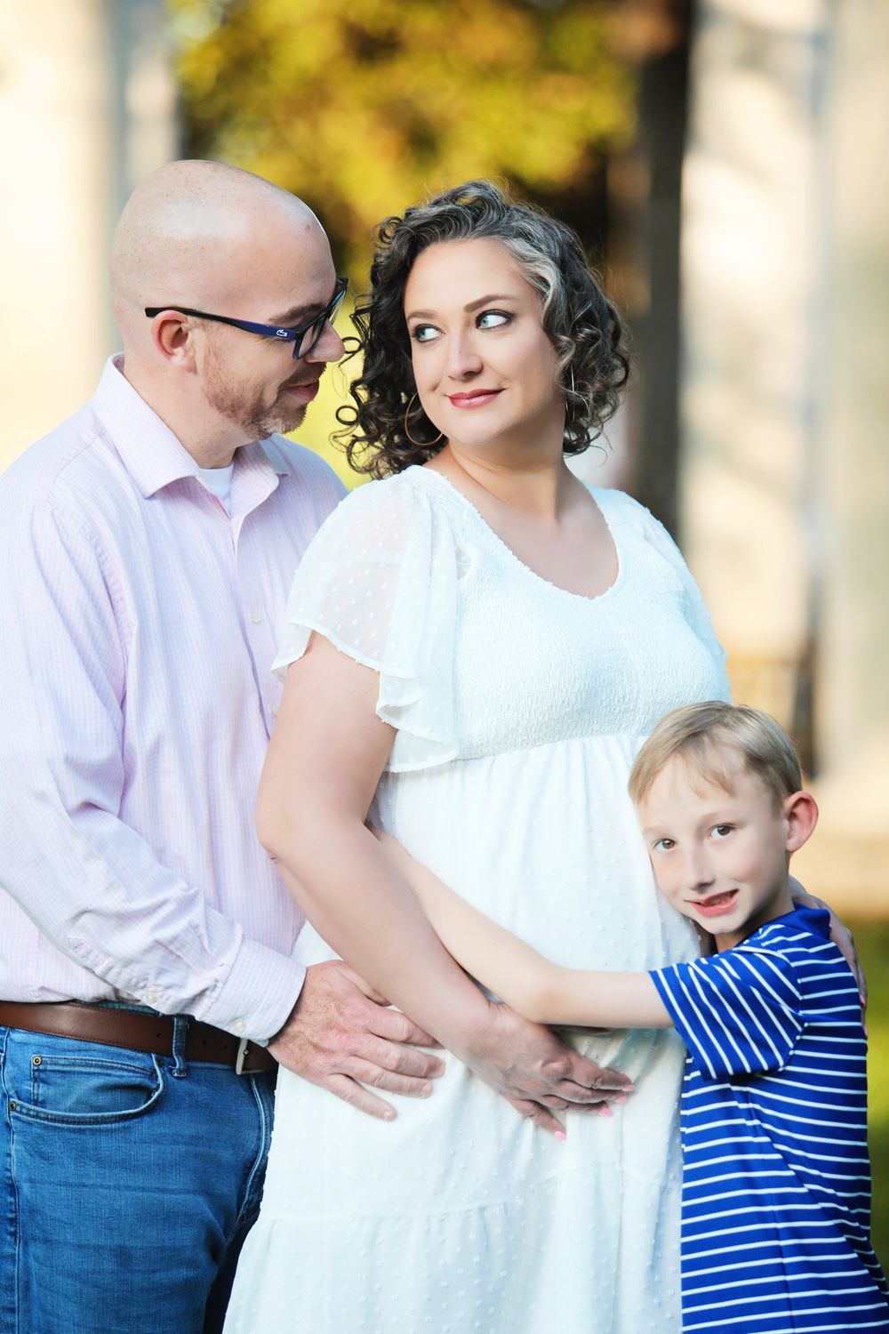 A family maternity portrait outdoors with autumn foliage in the background.