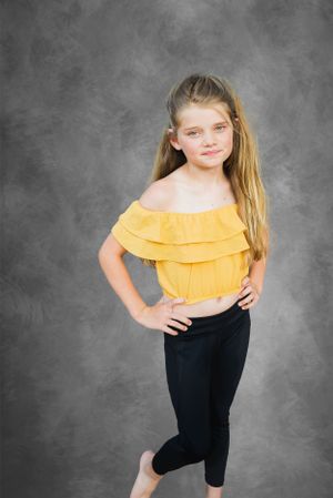 Young dancer in yellow off-shoulder top poses against gray backdrop.