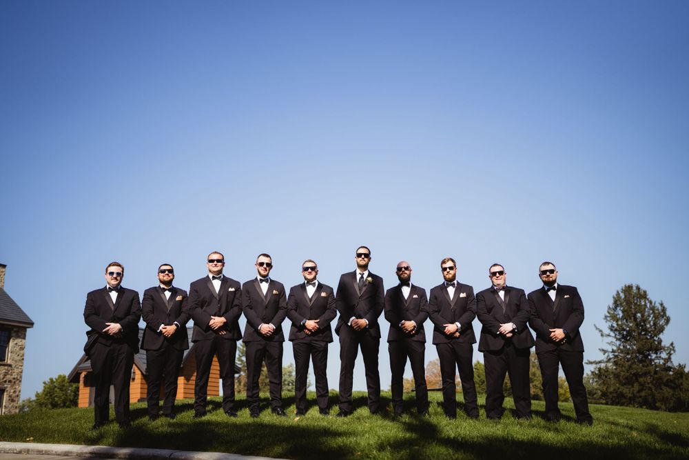 Row of groomsmen in black suits and bowties pose against blue sky backdrop.