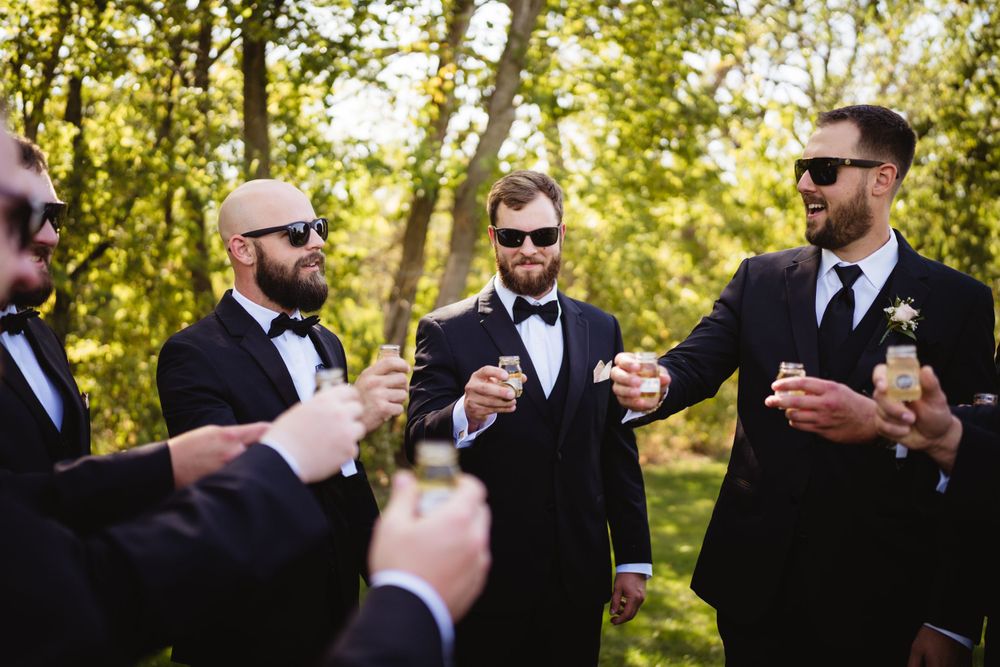 Groomsmen in black tuxedos toast with drinks during outdoor wedding celebration.
