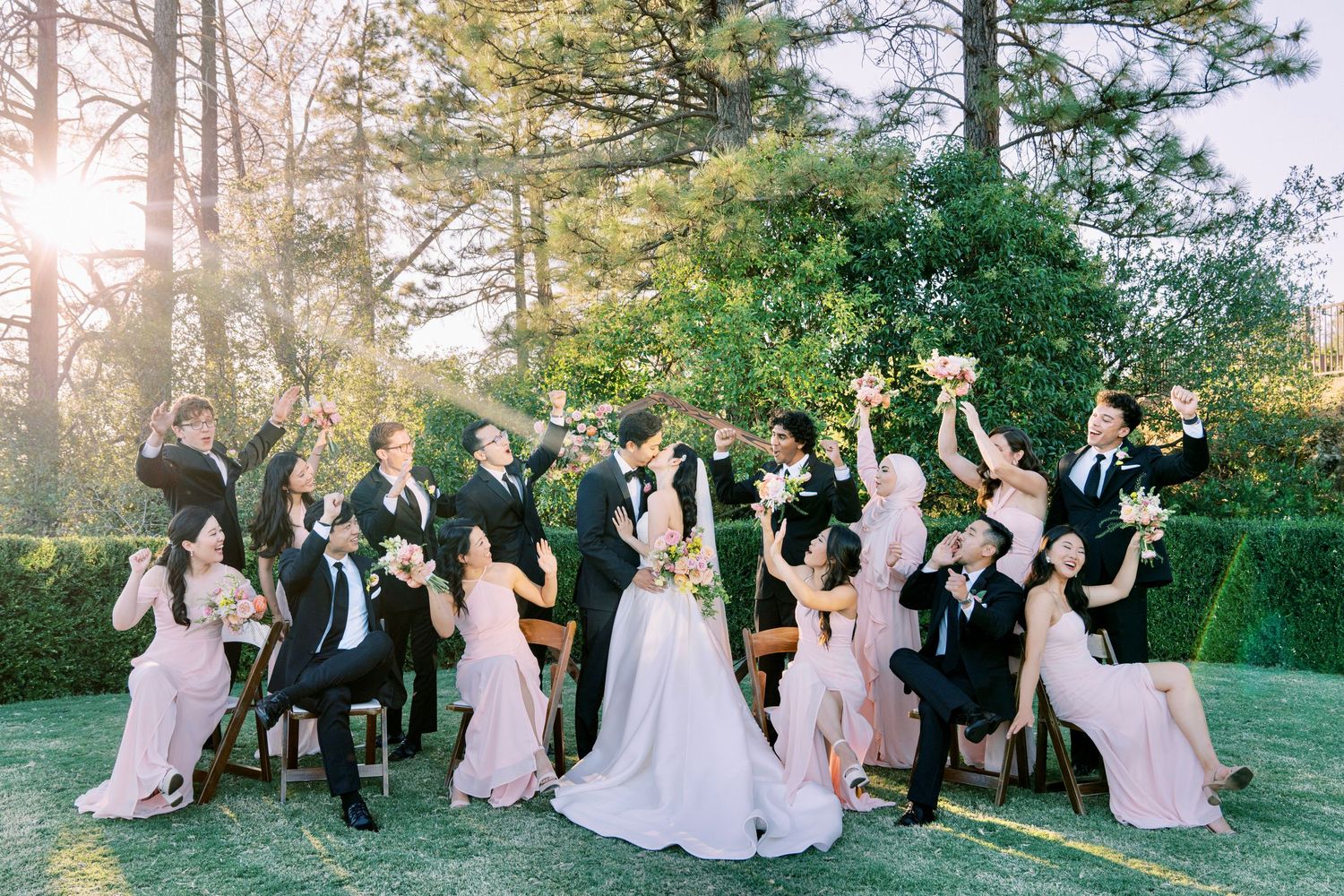 A joyful wedding party celebration on a sunny lawn surrounded by tall pine trees.