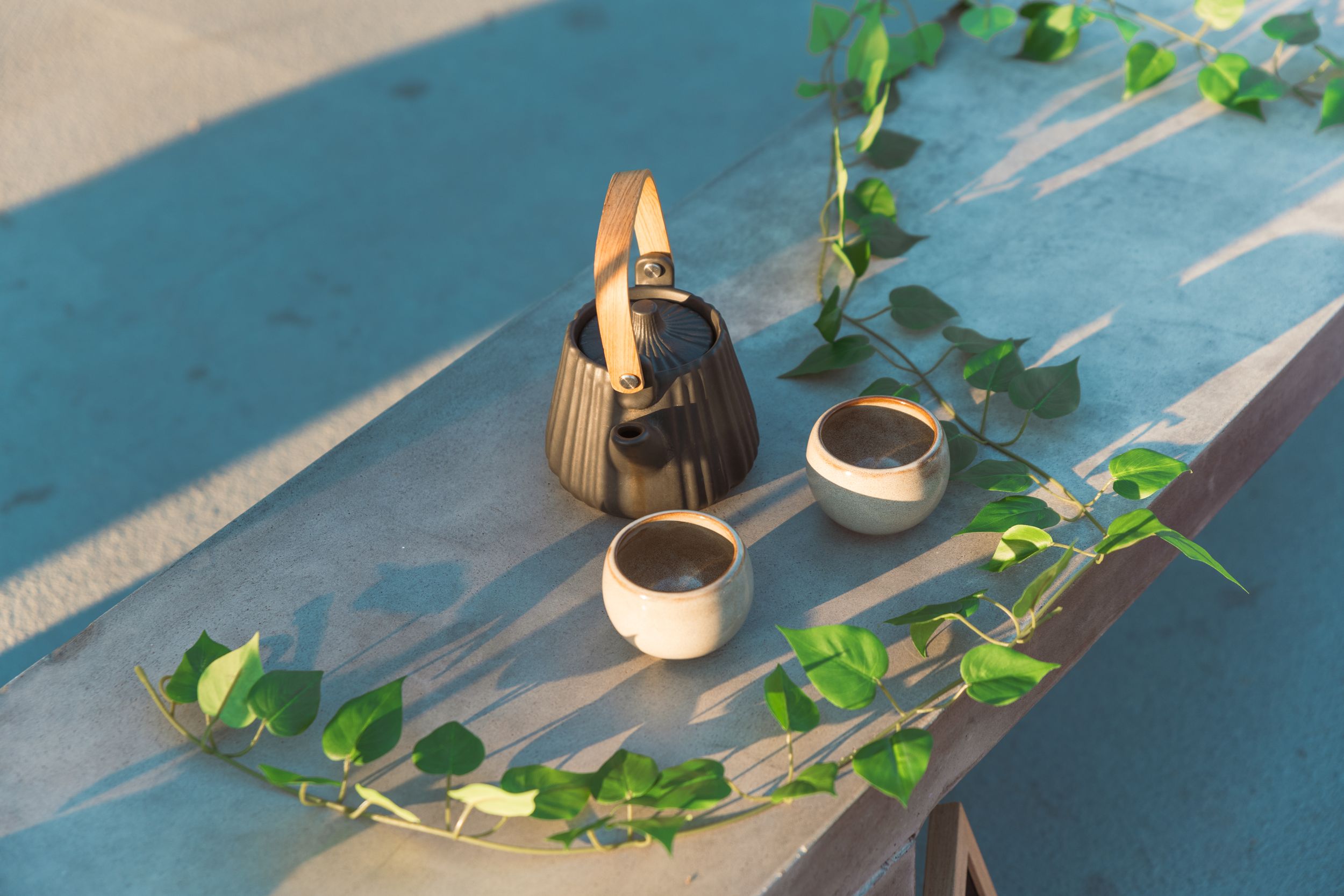 A trailing vine with bright green leaves lays on top of a stone bench and next to a ceramic teapot and traditional teacups