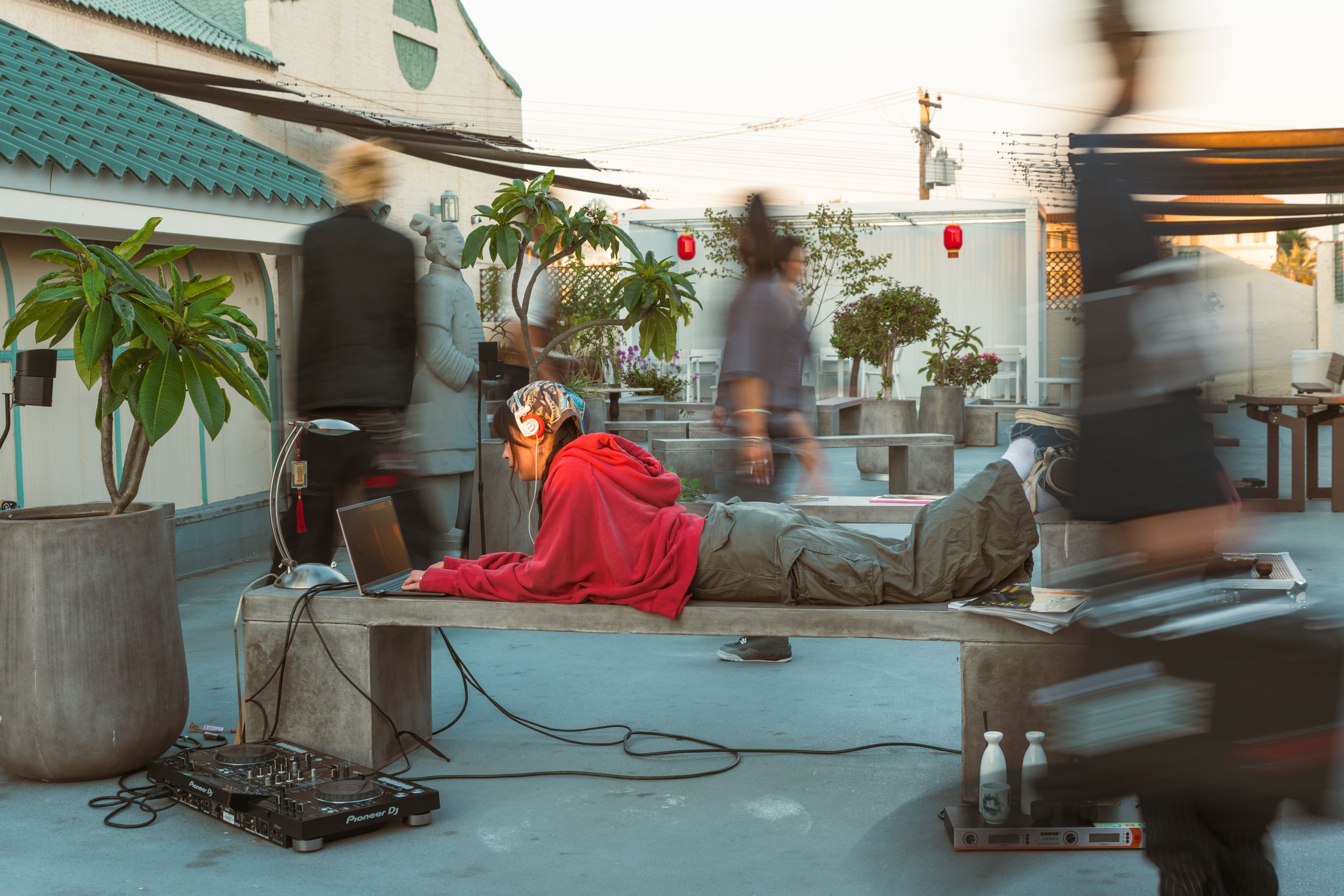 Person in a red outfit lies on a rooftop bench while blurred figures move past in an urban setting at dusk.