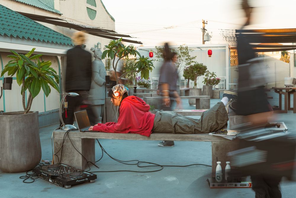 Person in a red outfit lies on a rooftop bench while blurred figures move past in an urban setting at dusk.
