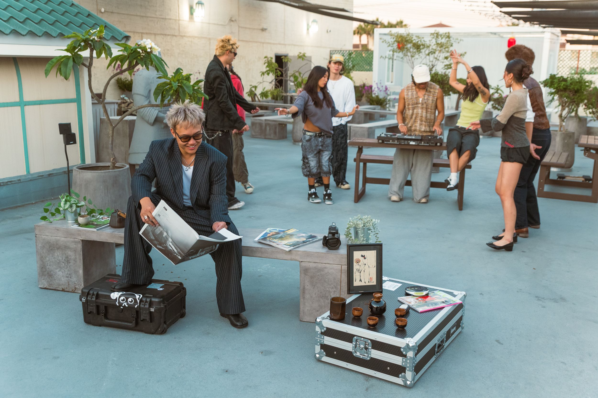 A man sits on an outdoor bench with one leg propped on a container while reading on a rooftop. People party in the background.