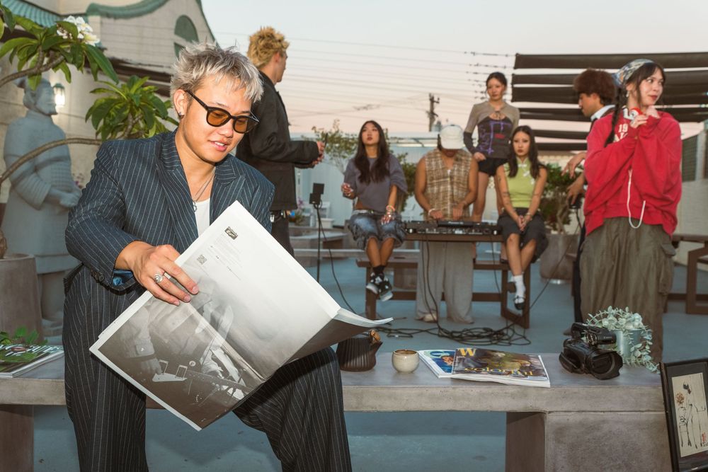 A group of people on a rooftop terrace while someone in sunglasses examines photographs during a casual gathering.