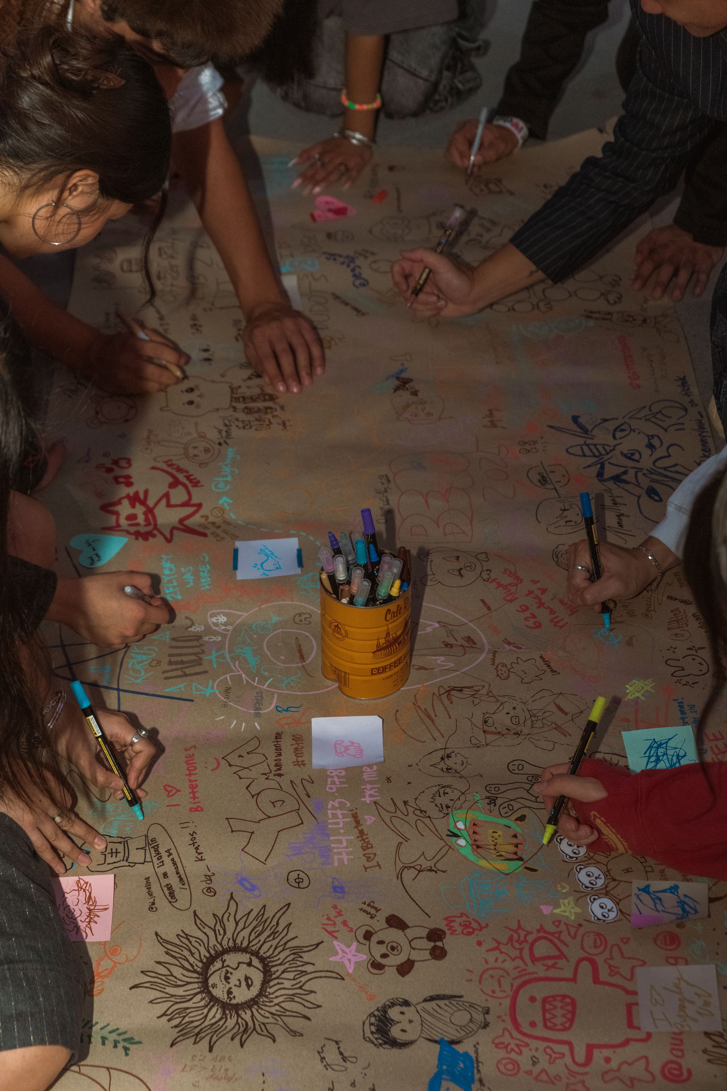 Hands and markers seen from above creating colorful drawings and designs on a large sheet of paper spread on a table.
