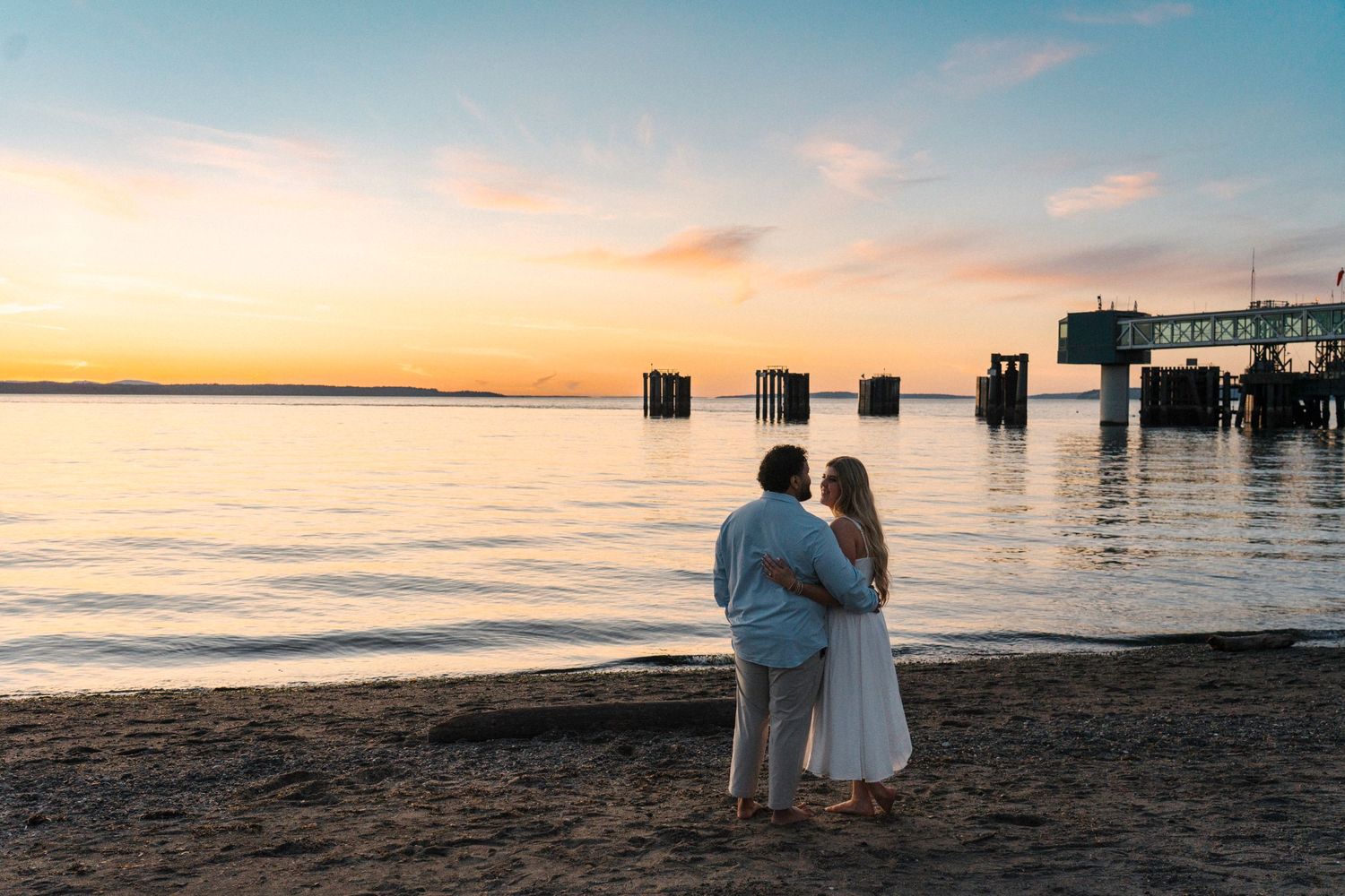 A couple embraces on a sandy beach at sunset with wooden pier pilings and calm water in the background.