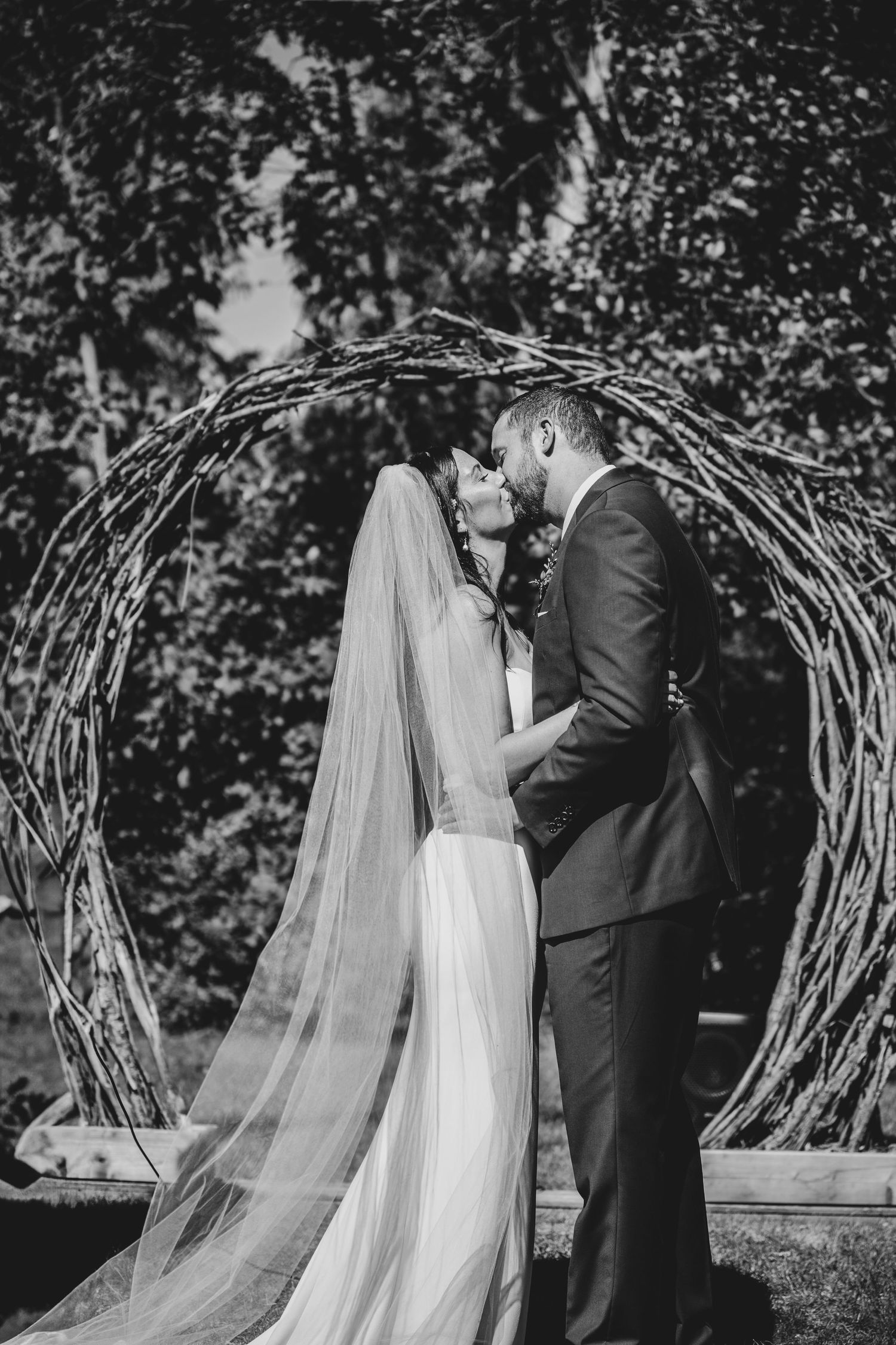 A romantic wedding scene shows a couple sharing a kiss under a rustic wooden arch in a garden setting.