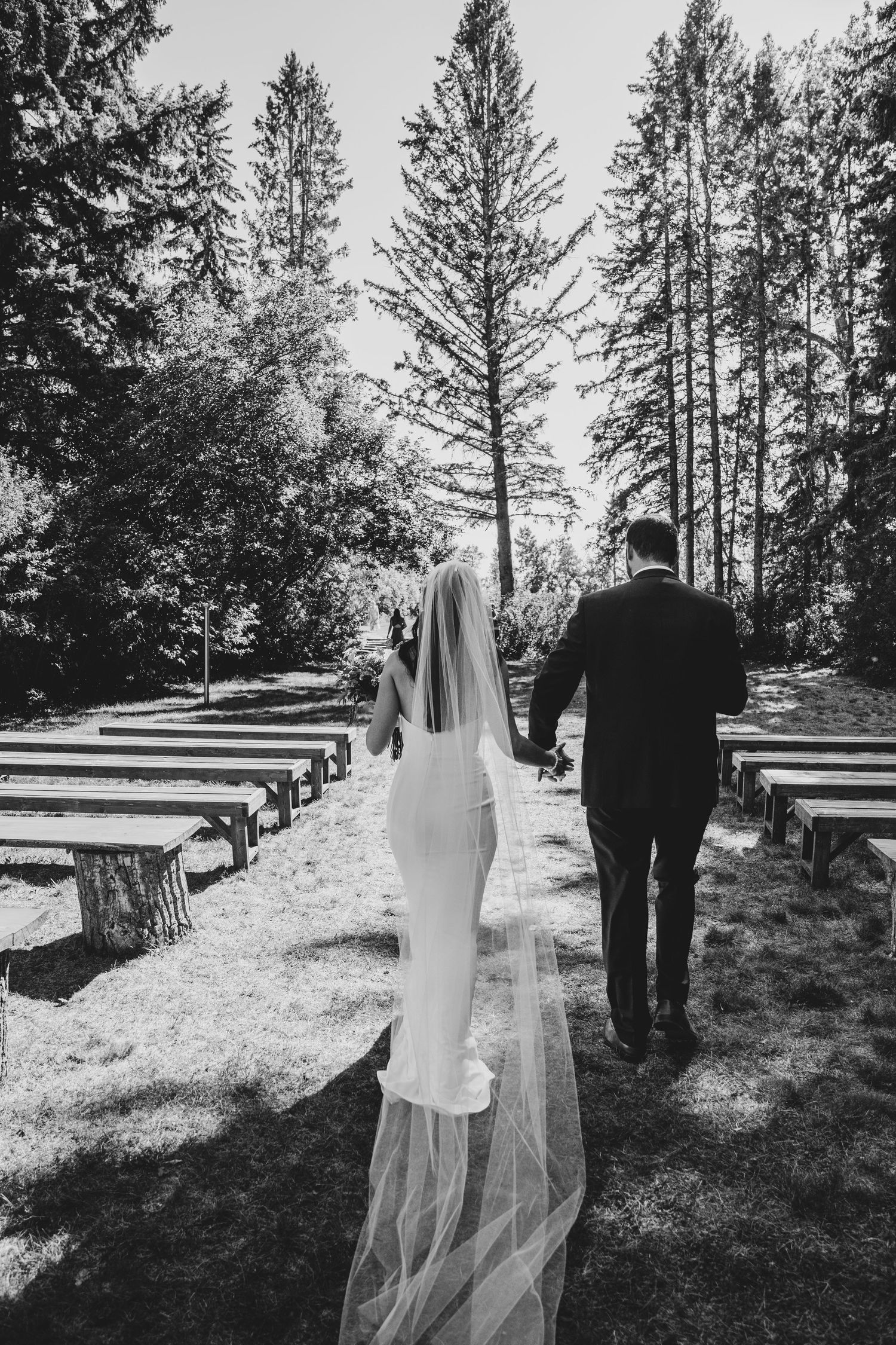 A couple walking down the aisle at an outdoor forest wedding venue with wooden benches and tall evergreen trees.