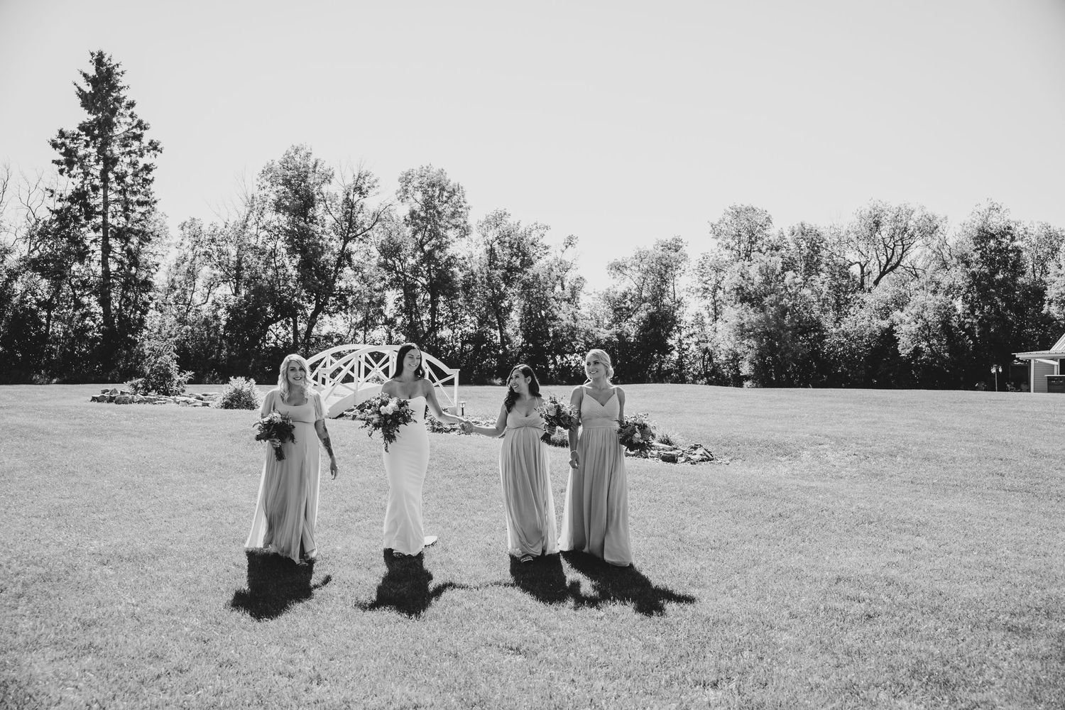 Four attendants in matching dresses dance and twirl together on a sunny lawn during a wedding celebration.