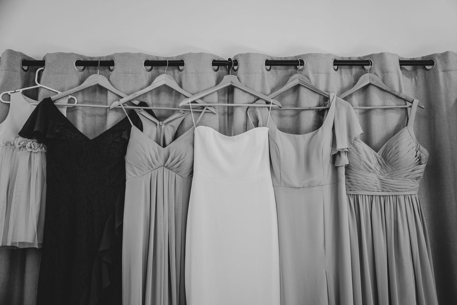 A row of bridesmaid dresses in various neutral tones hanging on wooden hangers against a light colored background.