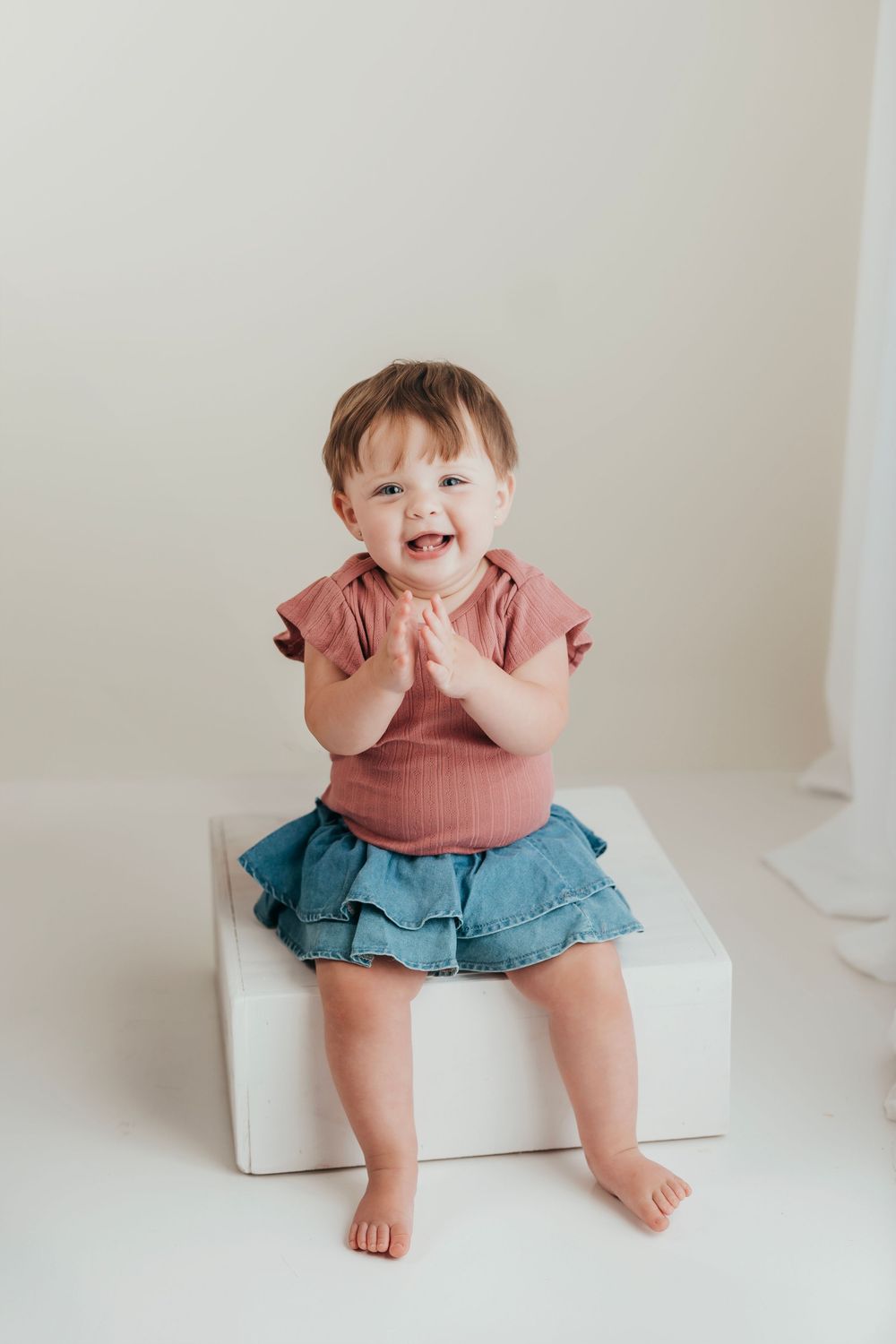 A happy toddler in a pink shirt and denim skirt sits and claps while perched on a white cube.