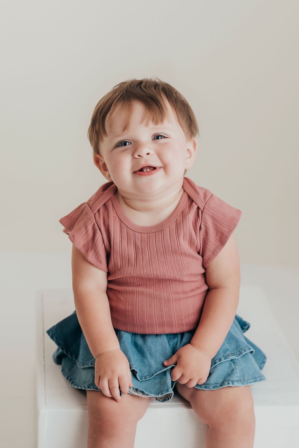 A happy toddler wearing a pink ruffled top and denim shorts sits on white background in a candid photo series.