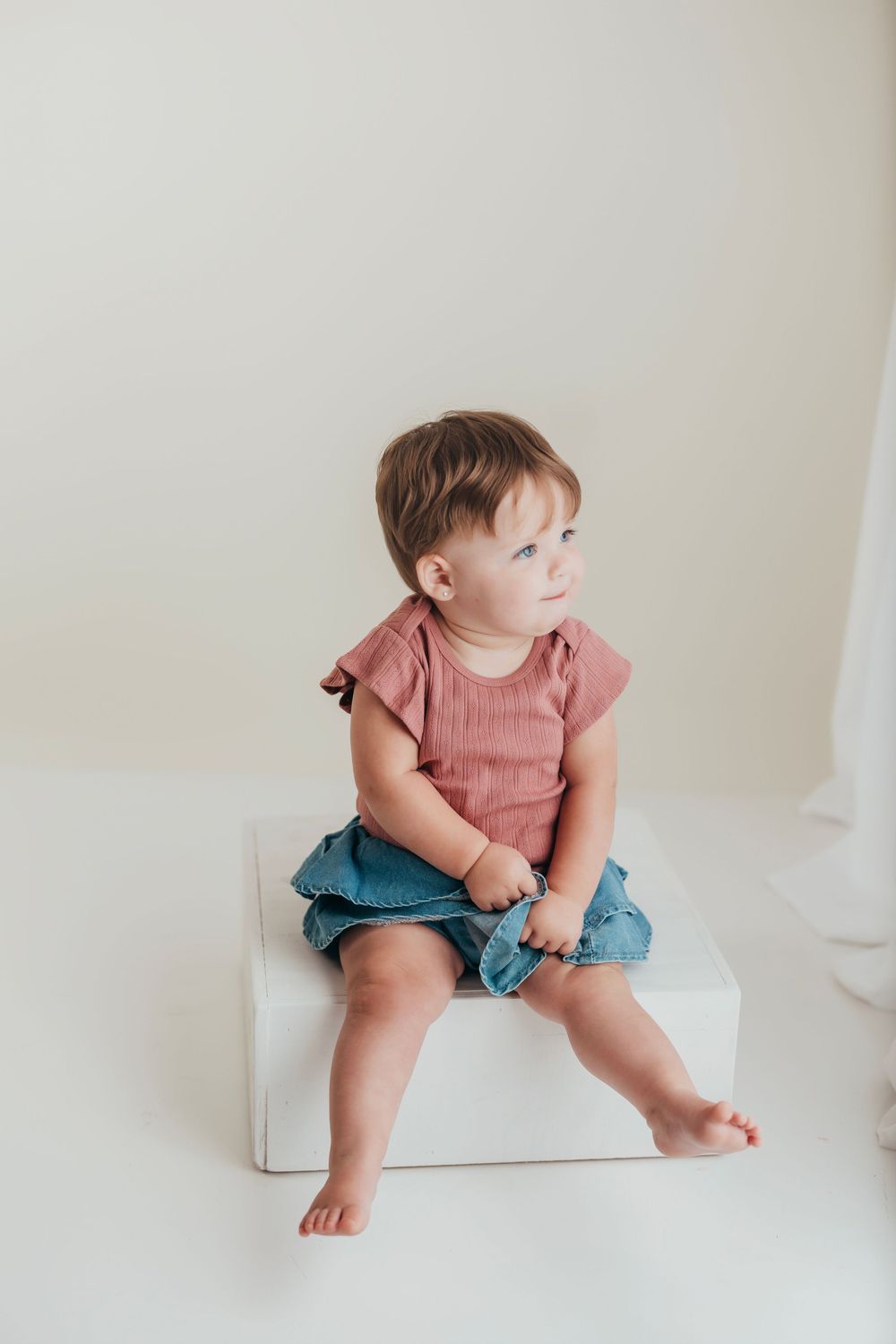 A toddler in a pink top and denim shorts sits and plays on a light-colored floor with a minimalist background.