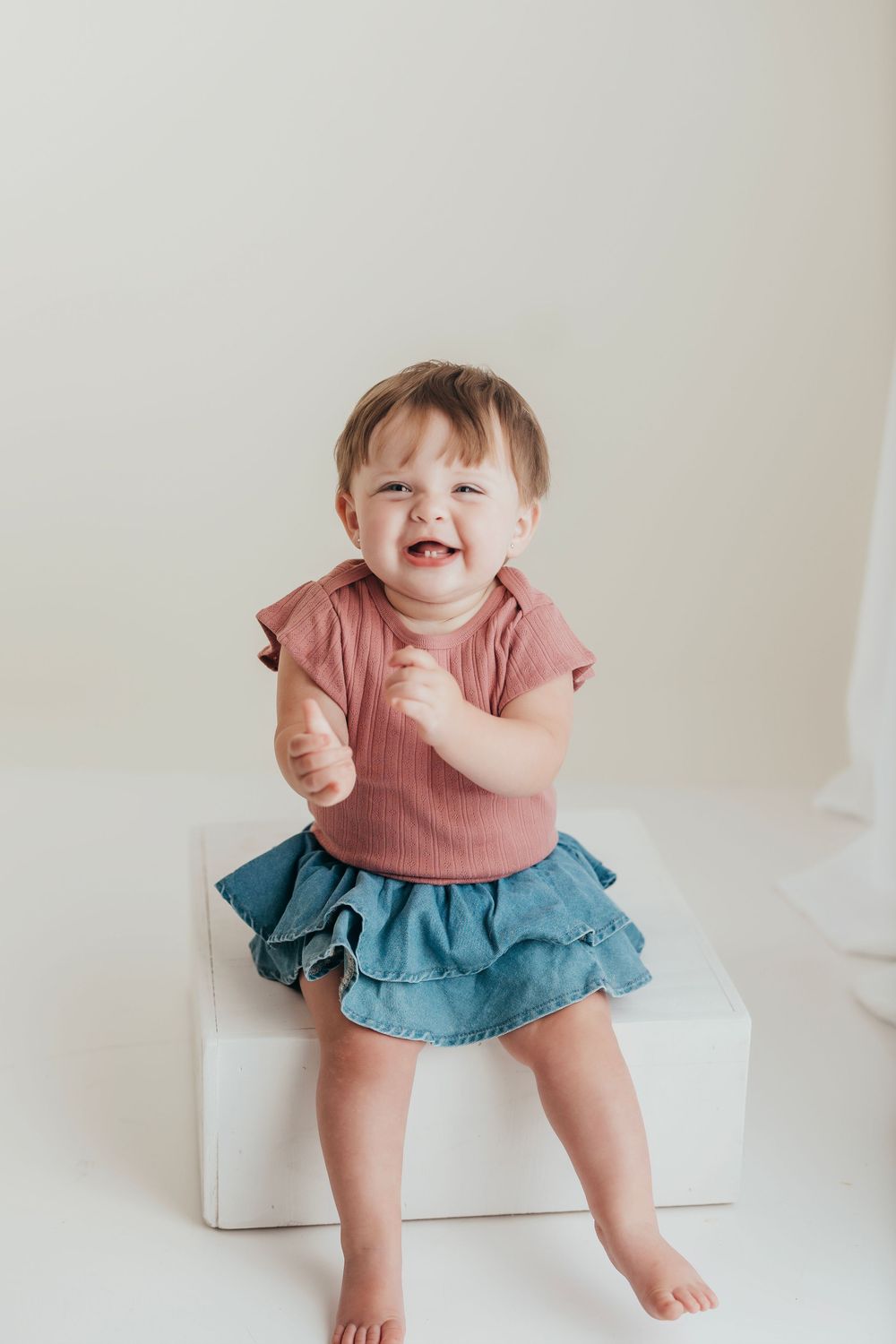 A happy baby wearing a pink top and blue ruffle skirt sits on a white cube against a neutral background.