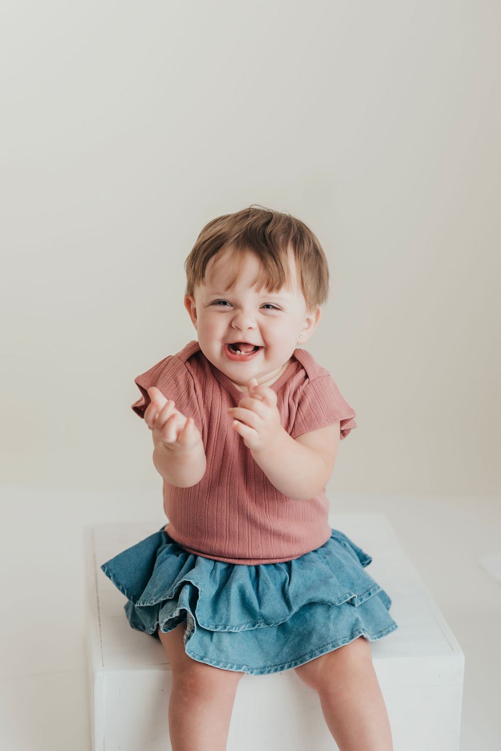 Small child sitting on white surface wearing pink ribbed shirt and blue ruffled skirt with joyful expression.
