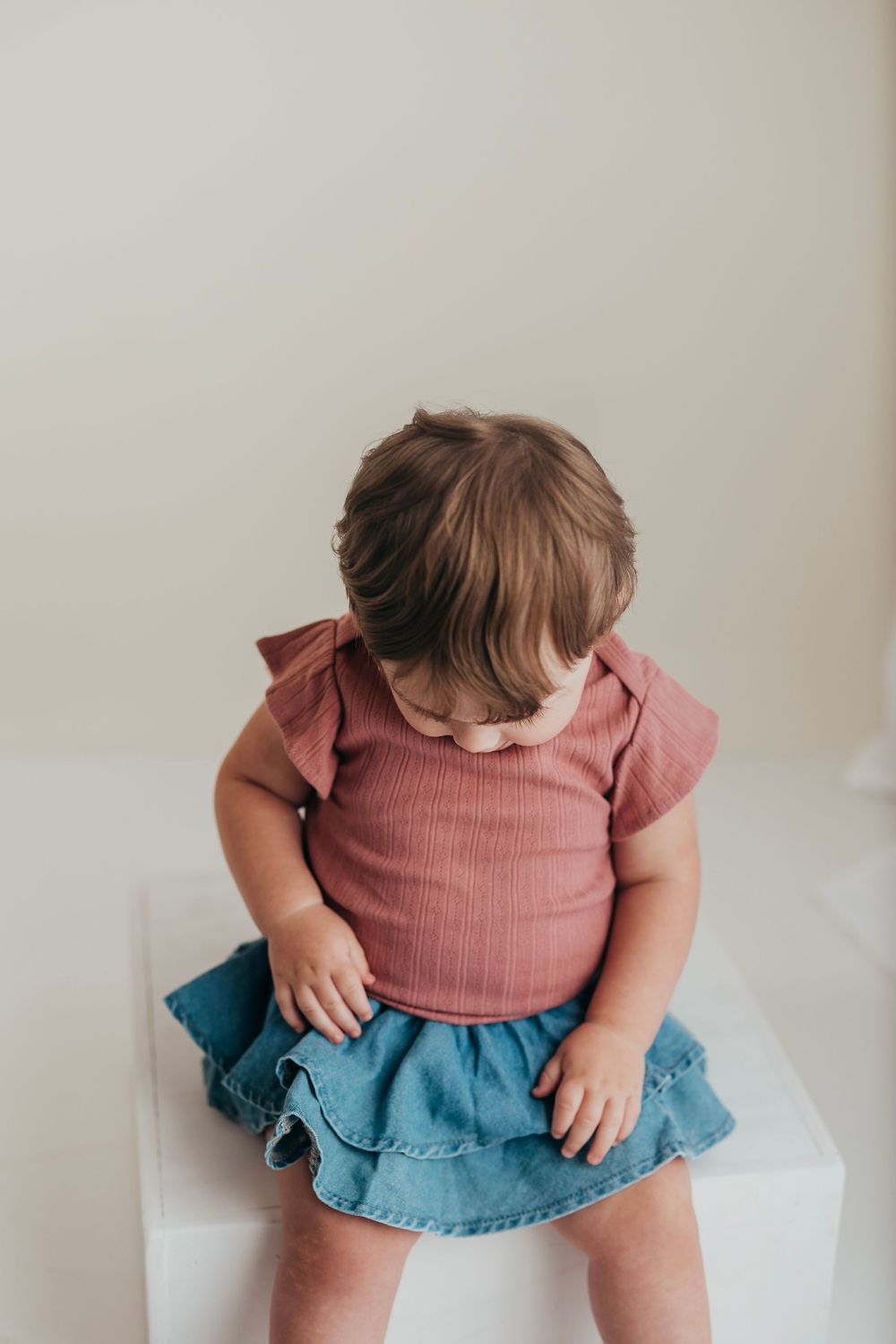 Small child wearing a pink ruffle-sleeve top and denim skirt looks down while sitting on white surface.