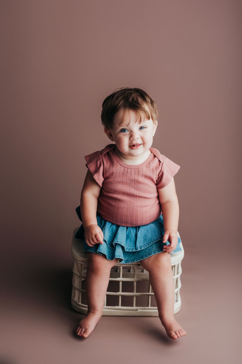 Toddler in pink top and blue skirt sitting on white stool against mauve backdrop.