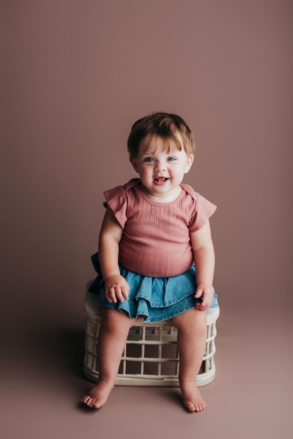 A baby in a pink top and denim skirt sits playfully on a vintage wooden bench against a mauve backdrop.