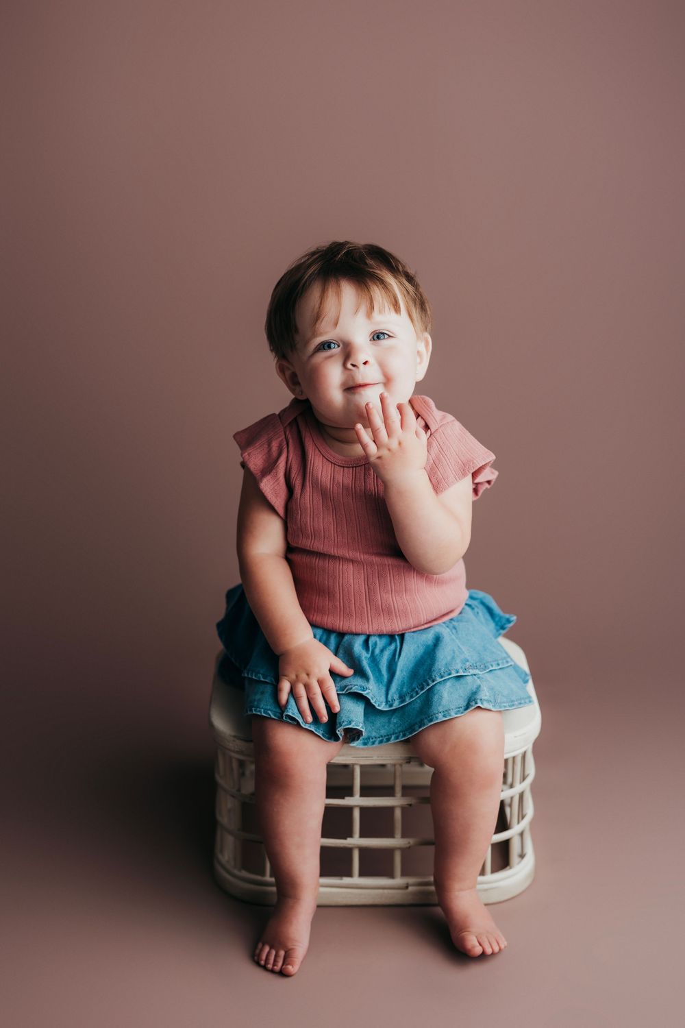 Baby in pink shirt and denim shorts sits on white crate against mauve background, touching face with hand.