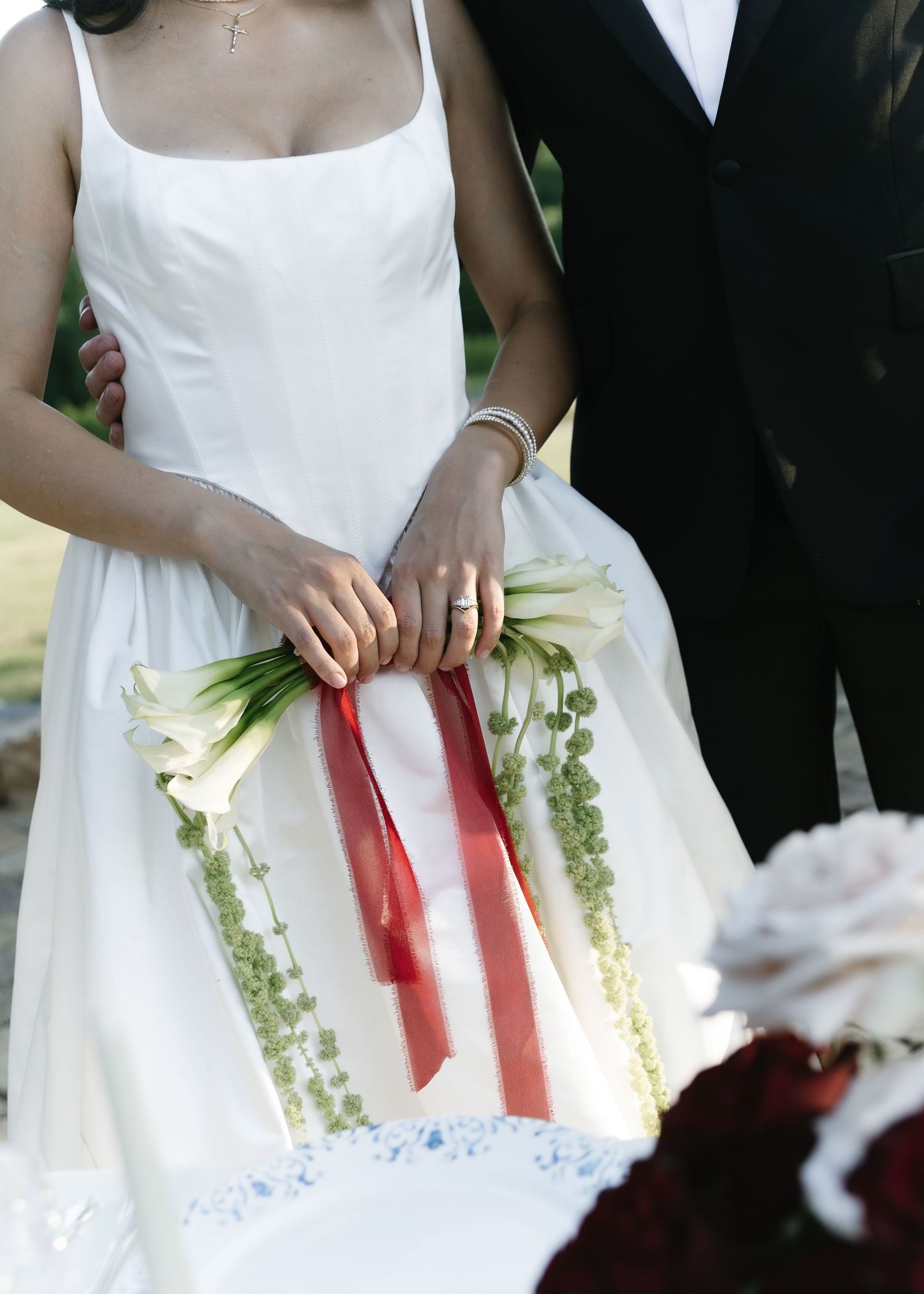 A wedding scene showing a bride in white dress holding a calla lily bouquet with red ribbons next to a person in black suit.