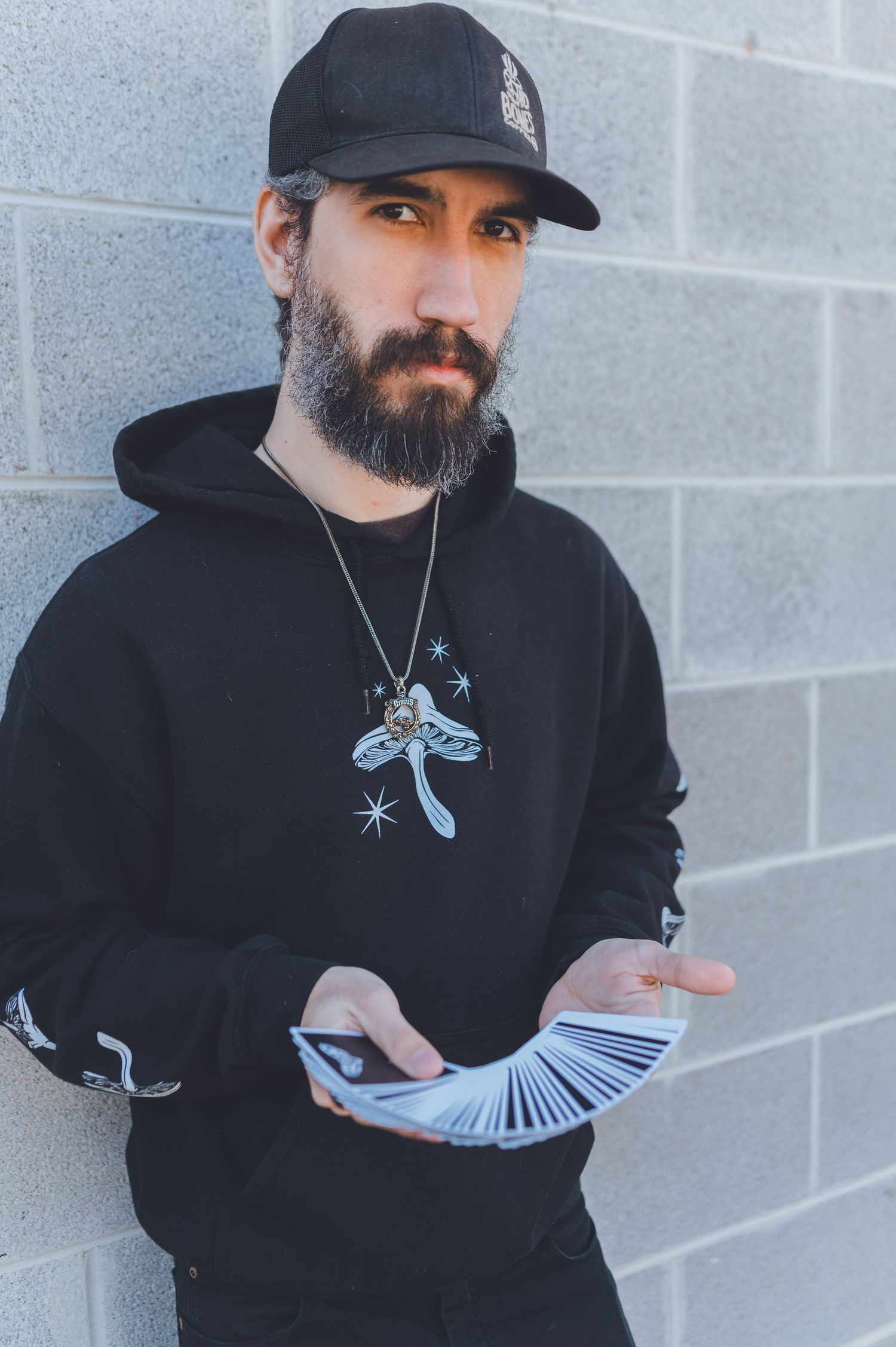 Person in black hoodie and baseball cap performs card flourish against gray wall while showing fanned deck.