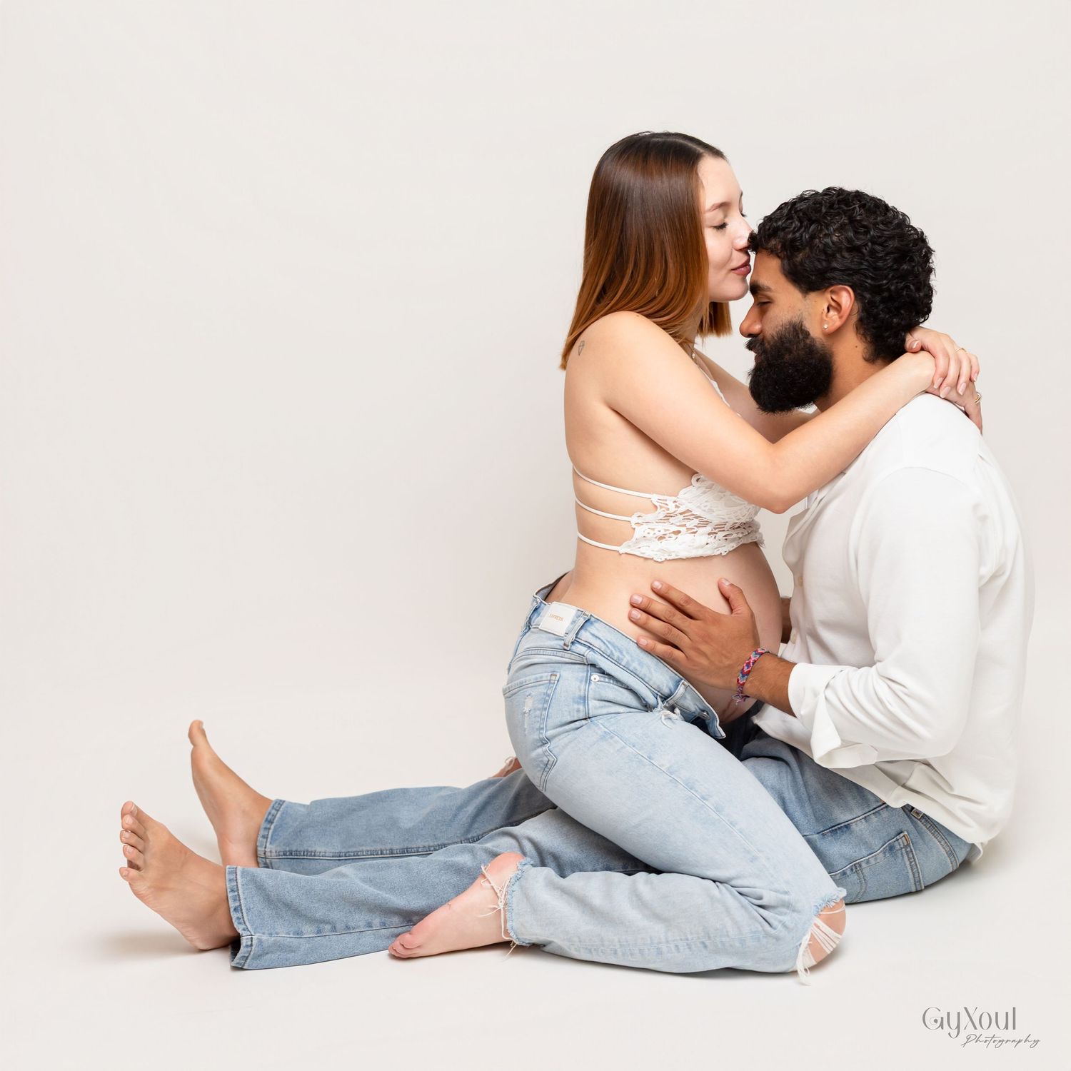 Intimate couple in casual attire embracing while sitting on white floor against neutral background.