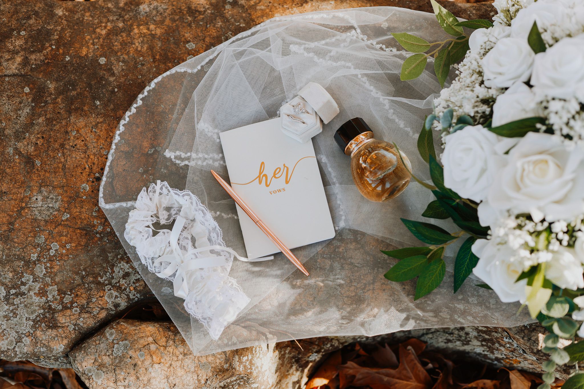 Wedding details flatlay with white roses, rings, and delicate lace elements on a rustic surface.