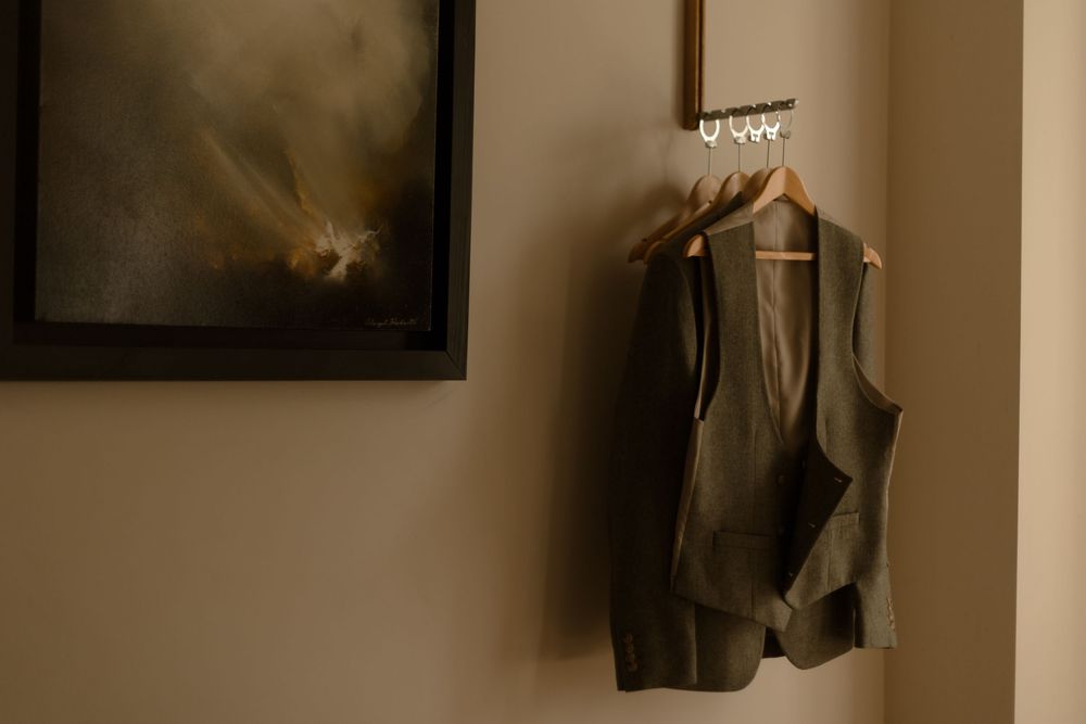 A green vintage dress vest hangs on a wooden hanger against a beige wall with framed artwork in the background.