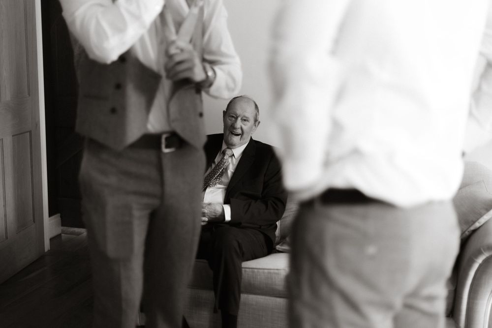 Black and white photograph showing someone seated against a wall between two standing figures in light colored clothing.
