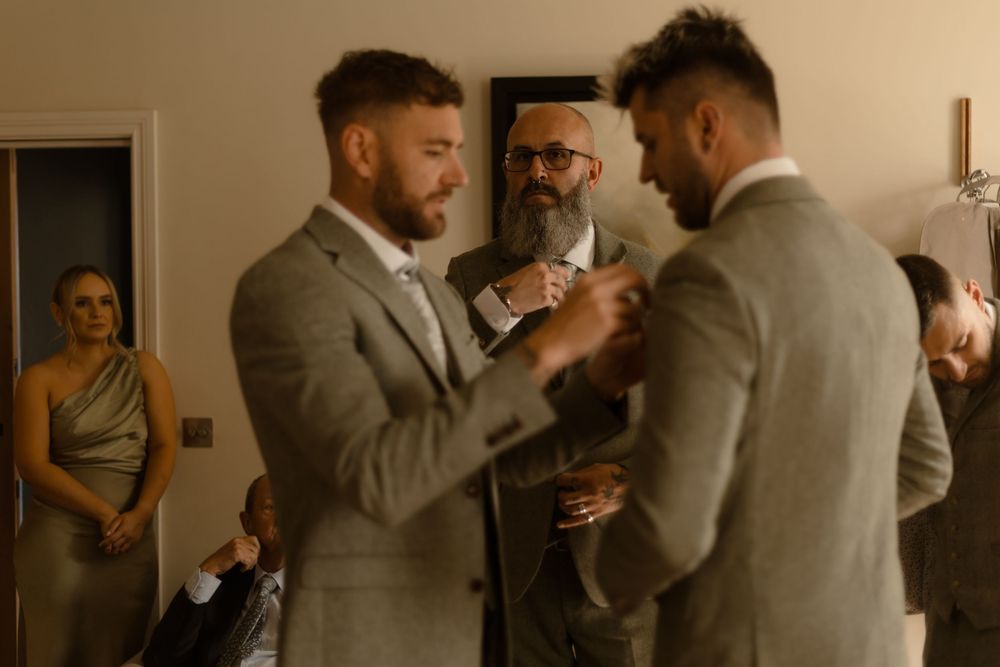 A warm moment between groomsmen in grey suits during a wedding preparation, with a guest watching in the background.