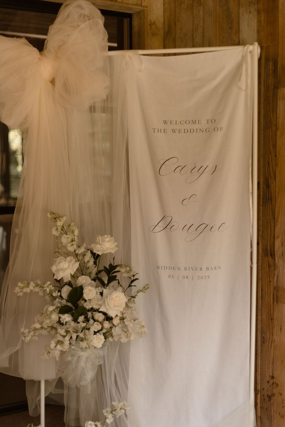Elegant wedding welcome sign on white fabric with calligraphy text and floral arrangement against wooden backdrop.