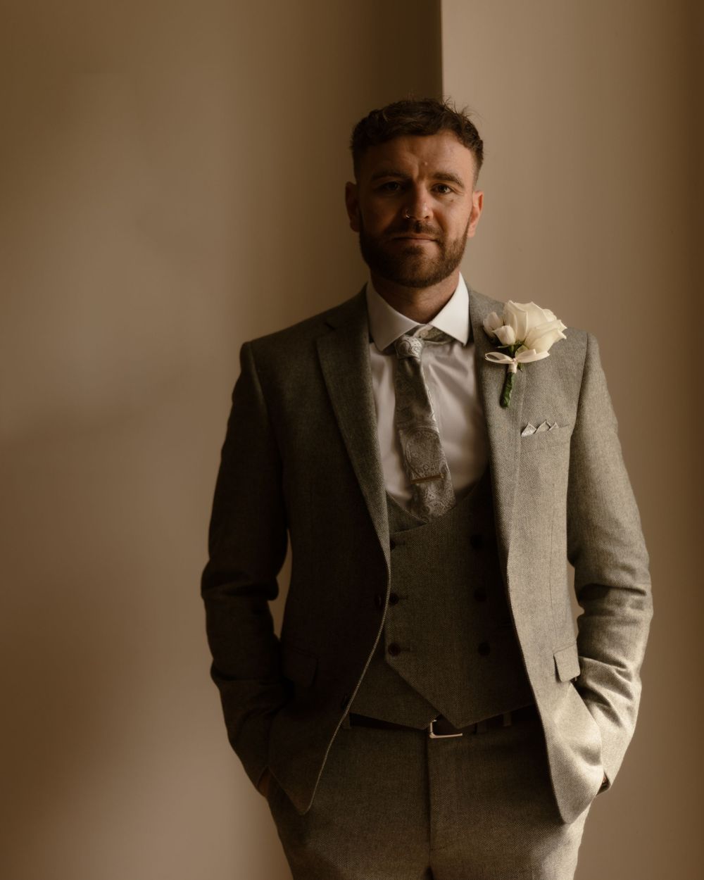 A groom in a light grey three-piece suit with a boutonniere poses with hands in pockets against a neutral wall.