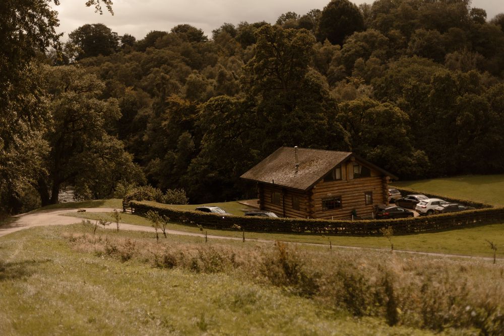 A rustic wooden cabin sits nestled against a forested hillside with parked cars and a winding gravel driveway.