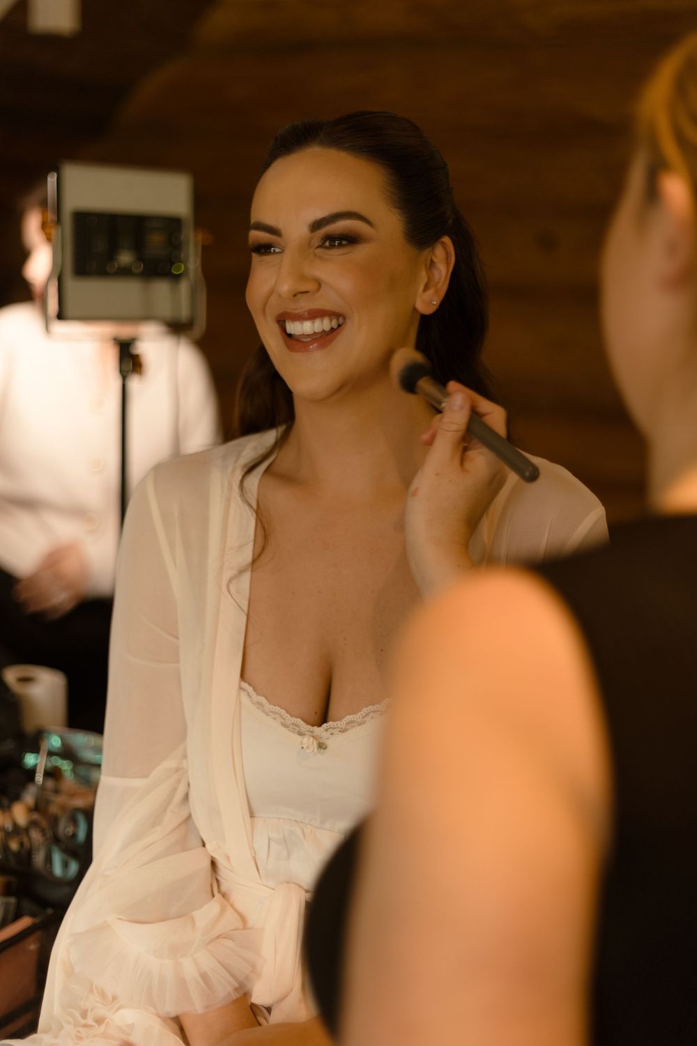 A bride smiles joyfully while getting ready on her wedding day, wearing a white robe and having makeup applied.
