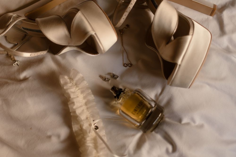 Wedding accessories including beige high heels and perfume bottle arranged on light fabric.