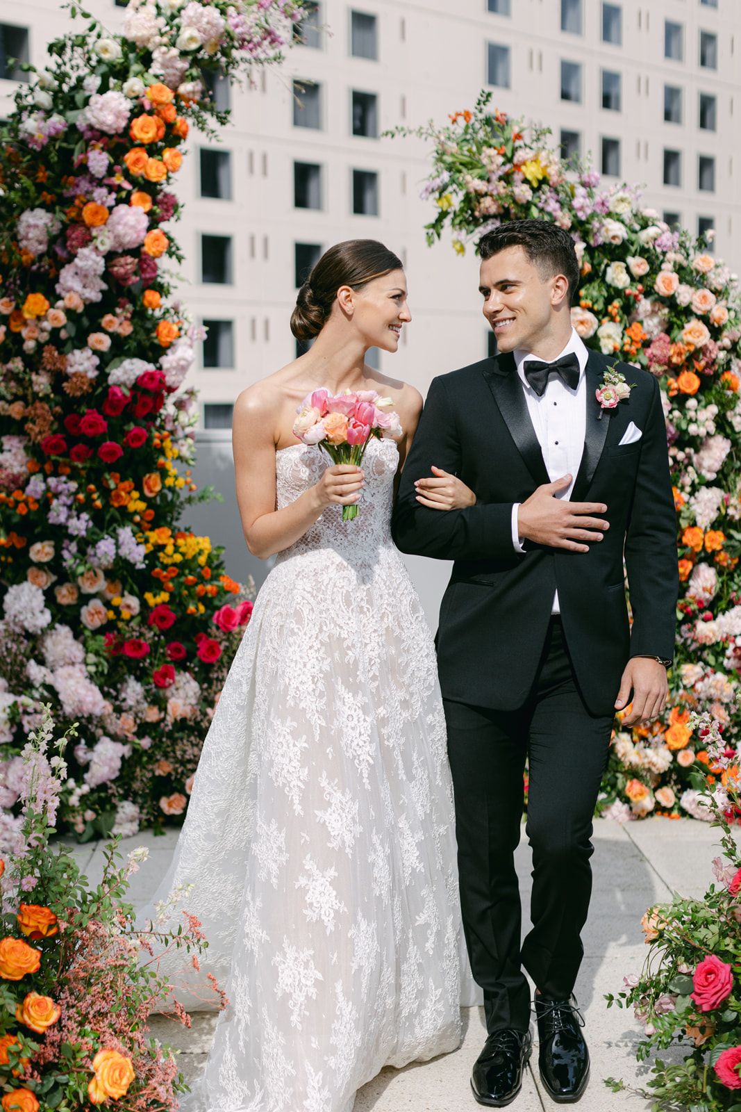 A beautiful outdoor wedding scene with a lush floral arch featuring orange and pink blooms against an urban backdrop.