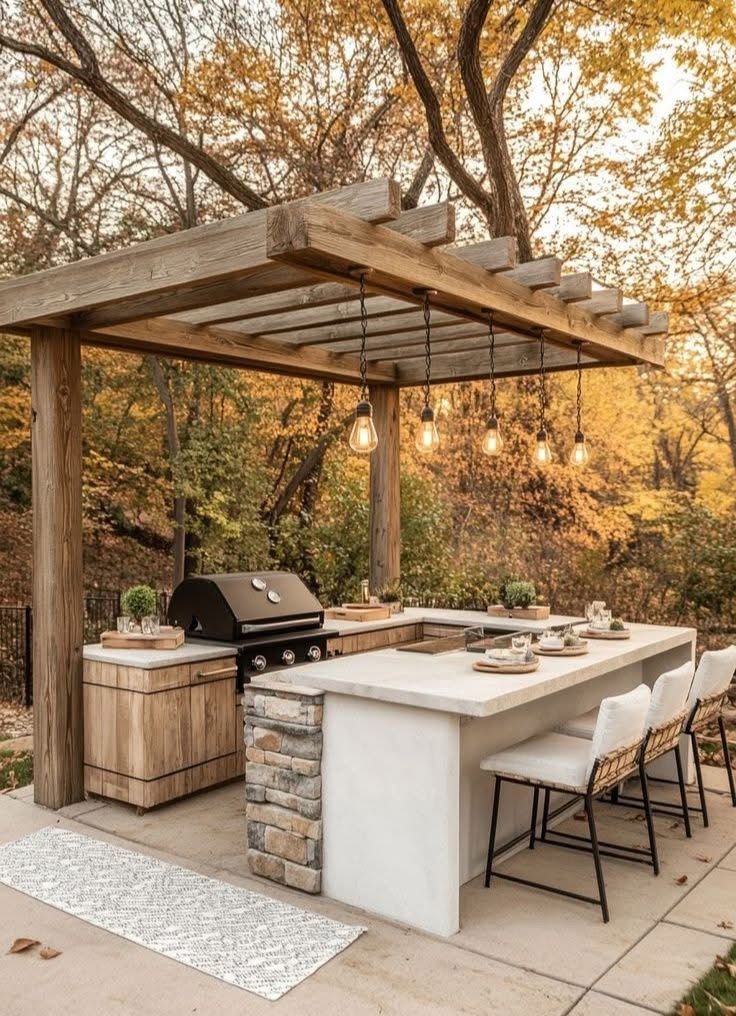 A modern outdoor kitchen with wooden pergola features stainless appliances and stone countertops at sunset.