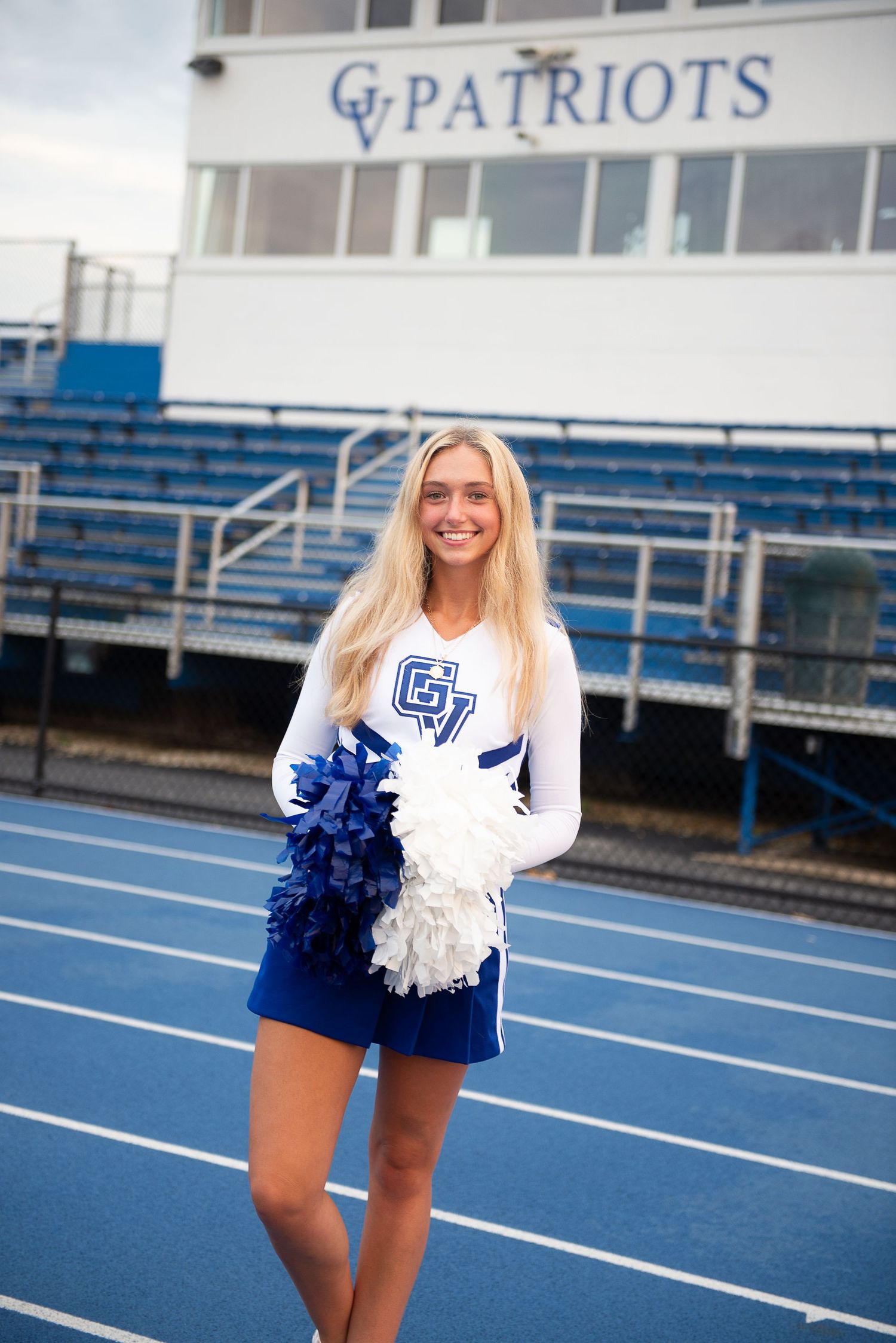 Cheerleader in white uniform and blue skirt performs on blue running track at football stadium.