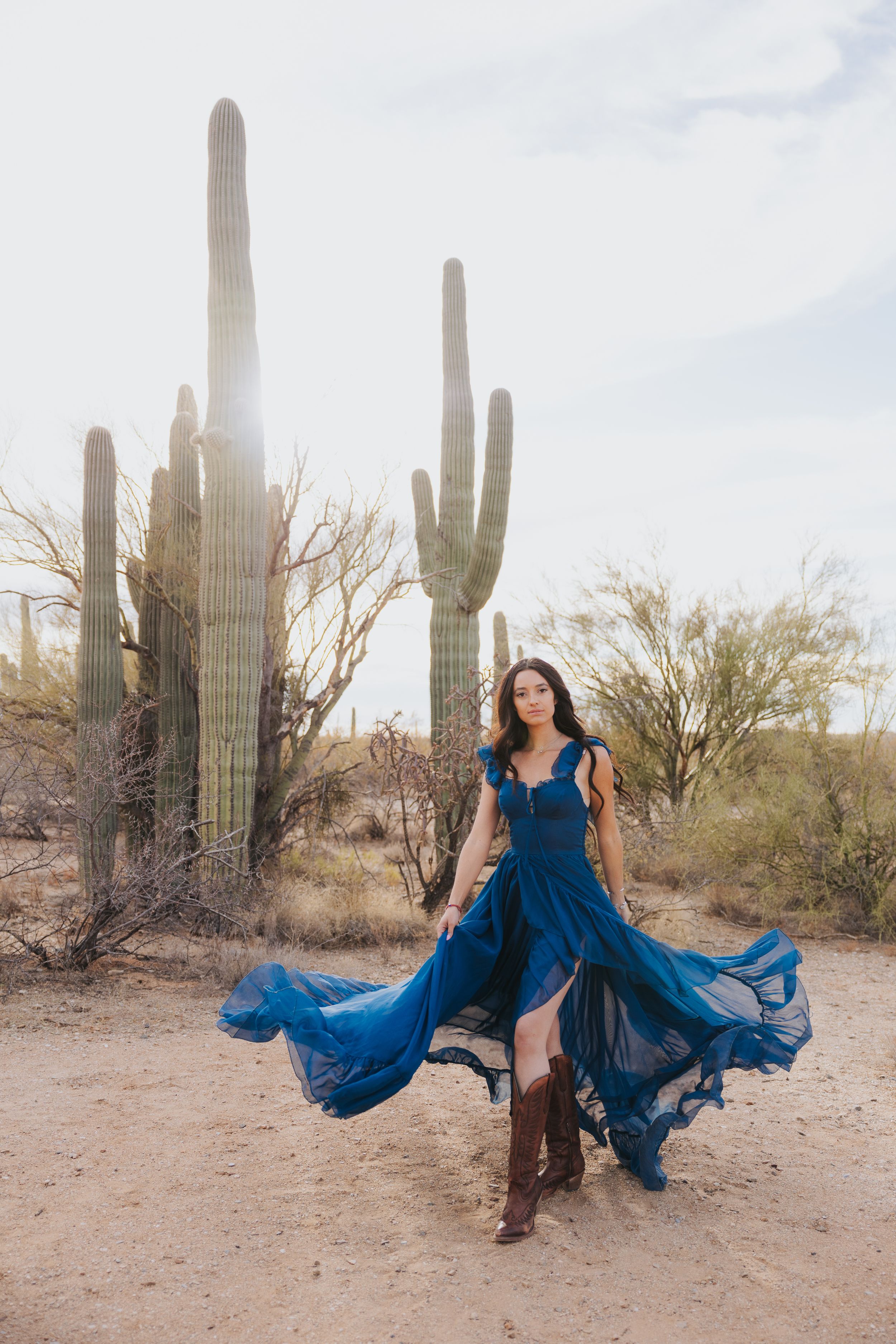 A flowing blue dress creates movement against tall saguaro cacti in the desert.