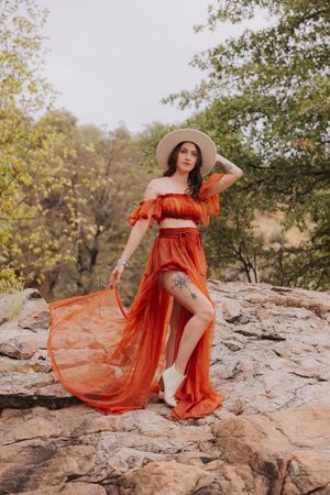 A flowing orange dress drapes over rocky terrain in a desert setting with trees in the background.