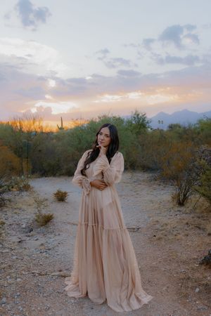 A soft pink dress flows in the desert sunset with mountains and saguaro cacti silhouetted against the sky.
